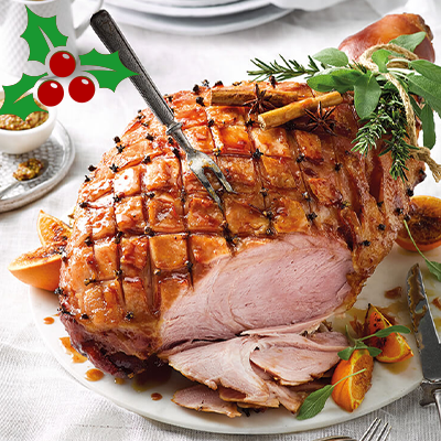 A sliced dressed Ham with cloves and garnishes sits on a white table cloth with a serving fork sticking out of it.