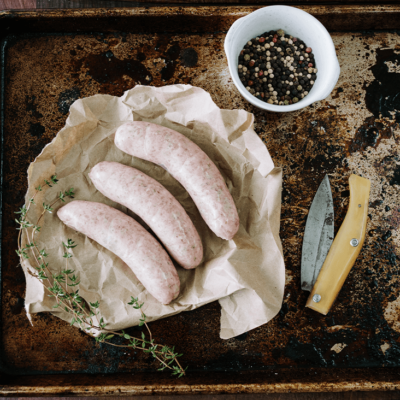 3 uncooked Old English sausages sit on a tray next to a knife and a small white bowl of peppercorns.