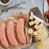 Overview shot of three uncooked sausages on a brown paper on a wooden board. Ingredients surround it, including olive oil, some grated and chunks of chedder cheese, a small white pot filled with pink salt and peppercorns and some cut up beef fillet.