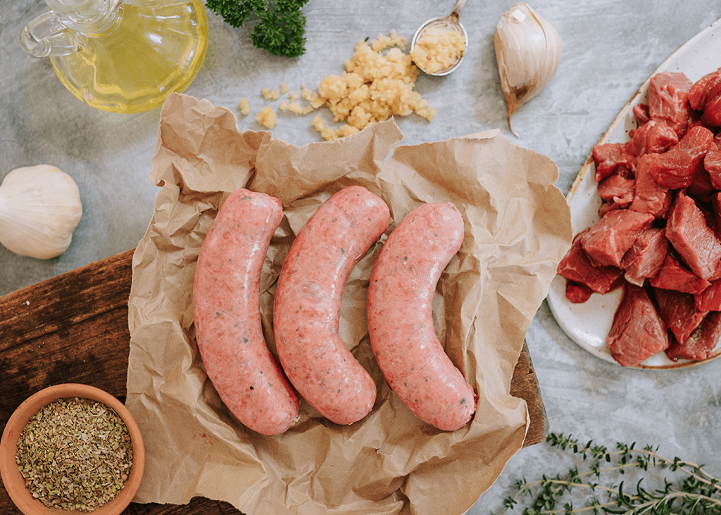 Overview shot of three uncooked sausages on a brown paper on a wooden board. Ingredients surround it, including olive oil, a pot of dried herbs, garlic cloves, some crushed garlic and some cut up beef fillet with some sprigs of thyme.
