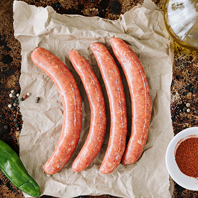 Four uncooked Lamb Merguez sausages sit on some paper on a roasting pan, bottom left is a chilli, bottom right is a small round white dish filled with paprika and a small glass bottle of olive oil sits at the top right of screen.