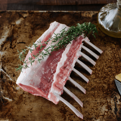A lamb rack lies on a tray with sprigs of fresh thyme on tope and a bottle of olive oil behind it.
