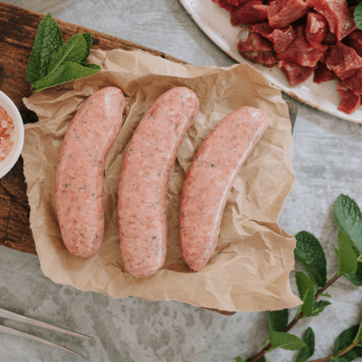 Overview shot of three uncooked sausages on a brown paper on a wooden board. Ingredients surround it, a pot of pink rock salt, fresh mint sprigs and some cut up lamb fillet with a small sprig of mint on top.