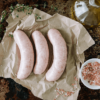 3 uncooked pork sausages sit on brown papers on a tray with thyme sprigs, a bottle of olive oil sits behind, with pink rock salt in a small white bowl in the front.