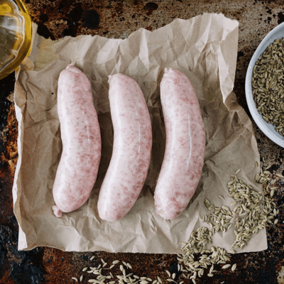 3 uncooked pork sausages sit on brown paper on the left is a small glass bottle of olive oil, on the right is a small white bowl of fennel seeds spilling over the brown paper.