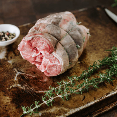 A boneless rolled lamb shoulder sits on a tray wit a small white bowl of peppercorns behind and 2 sprigs on rosemary sitting in front.