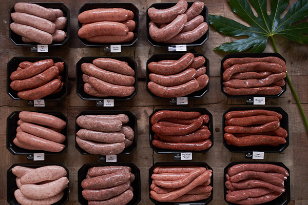 Overview of various sausage flavours packed in trays with a leaf garnish in the top right corner.