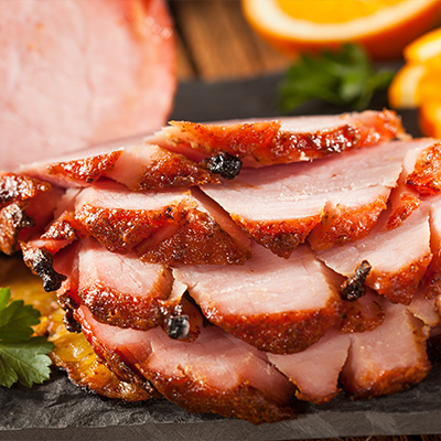 Stacked slices of Xmas baked ham with cloves sit in front of a baked ham surrounded by orange slices and a sprig of parsley.