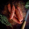 A group of kransky sausages are piled together, herbs and a knife sit either side of them.
