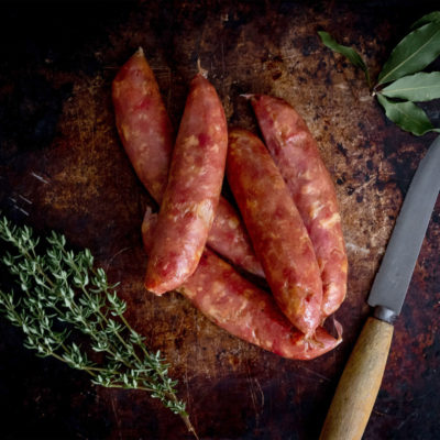 A group of kransky sausages are piled together, herbs and a knife sit either side of them.