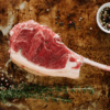 A Waipawa Butchery beef tomahawk steak sits on an oven tray with sprigs of herbs in front and a small white bowl of coloured peppercorns behind.
