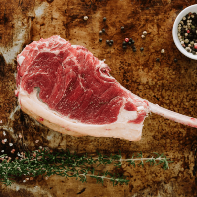 A Waipawa Butchery beef tomahawk steak sits on an oven tray with sprigs of herbs in front and a small white bowl of coloured peppercorns behind.