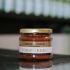 A jar of Maison Therese Tomato Relish sits on a reflective bench with out of focus bottles behind it.
