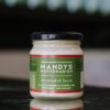 A jar of Mandys Horseradish sits on a reflective bench with out of focus bottles behind it