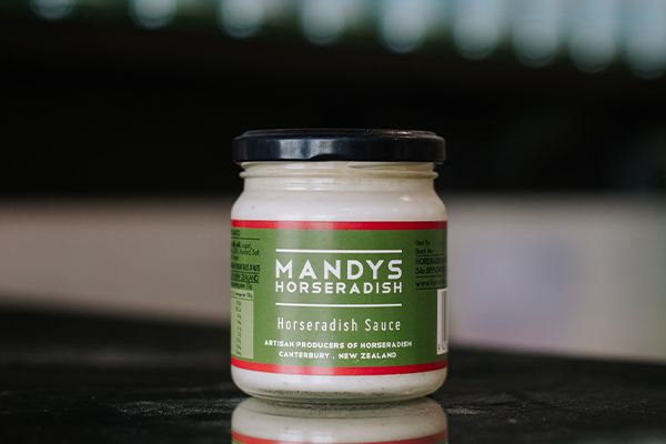 A jar of Mandys Horseradish sits on a reflective bench with out of focus bottles behind it