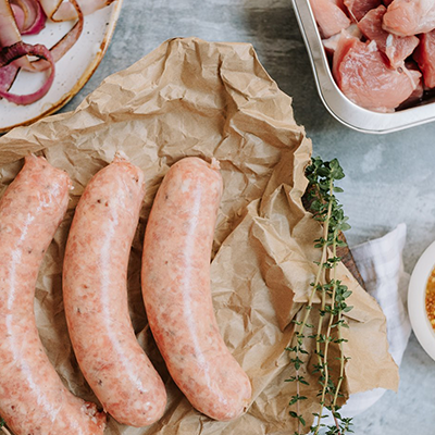 3 pork sausages sit on some crumpled brown paper. They are surrounded by a bowl of red onions, a small bowl of seeded mustard and a tray of pork pieces.