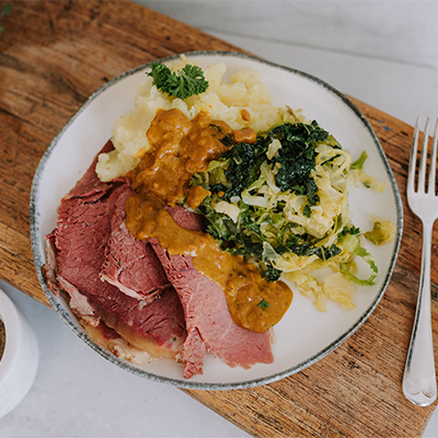 Waipawa Butchery Kitchen corned beef, mustard sauce, buttery cabbage and mashed potato on a white plate, sitting on a chopping board with a knife and fork and a white ceramic pot of black pepper with a spoon in the foreground.