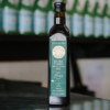 A bottle of The Village Press Avocado Oil sits on a reflective bench with out of focus bottles behind it.