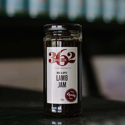 A jar of 362 Grill house lamb jam sits on a reflective bench with some out of focus bottles in the background.