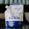 A pouch of little bone broth grass fed beef broth sits on a reflective bench with out of focus bottles behind it.