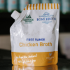 A pouch of little bone broth free range chicken broth sits on a reflective bench with out of focus bottles behind it.