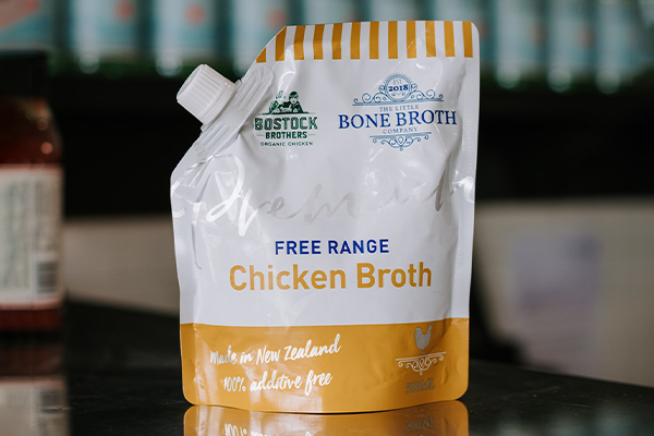 A pouch of little bone broth free range chicken broth sits on a reflective bench with out of focus bottles behind it.
