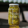 jar of Pickle and Pie Bread and Butter PIckle sits on a reflective bench with out of focus bottles behind it.