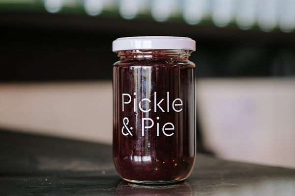 A jar of Pickle and Pie Cranberry Chutney sits on a reflective bench with out of focus bottles behind it.
