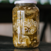 A jar of Pickle and Pie Dill Cucumbers sits on a reflective bench with a blurred background.