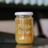 A jar of Pickle and Pie Hot Dog Relish sits on a reflective bench with out of focus bottles behind it.