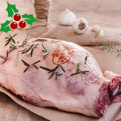 An uncooked leg of lamb sits on some brown paper on a wooden board. A sprig of rosemary sits on the lamb, behind you can see some garlic bulbs and brown string.