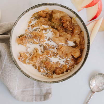 A bowl of apple crumble sits on a town next to a whole apple, a cut up apple and a dessert spoon.