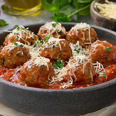 A dish of meatballs in a tomato sauce, topped with grated parmesan and herbs sits on a bench top surrounded by ingredients.