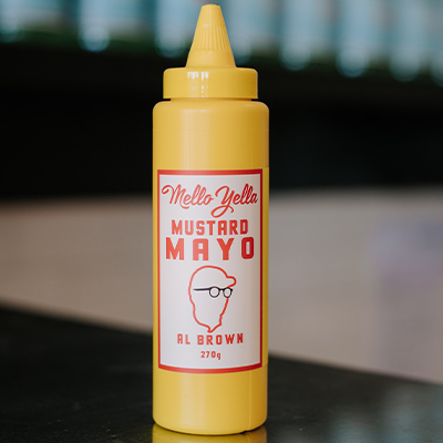 A bottle of Al Brown's mello yella mustard mayo sits on a reflective bench with some out of focus bottles in the background.