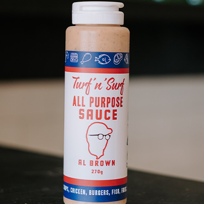 A bottle of Al Brown turf 'n' surf all purpose sauce sits on a reflective bench with some out of focus bottles in the background.