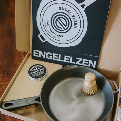 An open box with an Engelelzen fry pan with bamboo scrubber and wax sits on w wooden board.