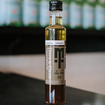 A jar of Maison Therese Date and Cider Vinaigrette sits on a reflective bench with out of focus bottles behind it.