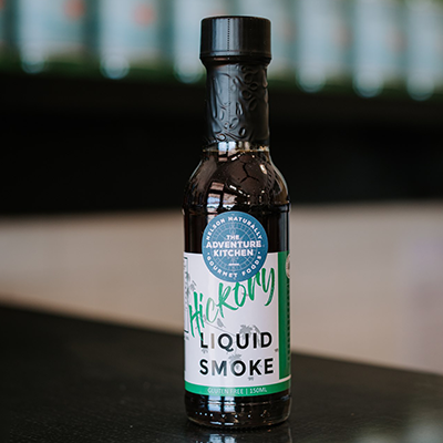 A bottle of The Adventure Kitchen Hickory Liquid Smoke sits on a bench with a blurred background.