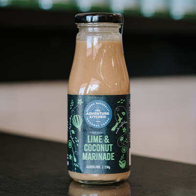 A bottle of The Adventure Kitchen Lime and Coconut Marinade sits on a bench with a blurred background.