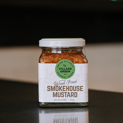 A glass jar of The Village Green Wood Fired Smokehouse Mustard sits on a bench with a blurred background.