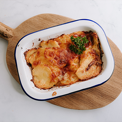 A dish of freshly baked potato gratin with a crispy golden top sits in a white dish on top of a wooden board with a handle.