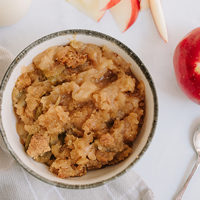 A white bowl of apple cruble sits on a shite table cloth next to a dessert spoon and crunchy red apple.